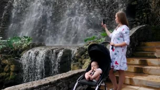 滝の近くの彼女の赤ちゃんを連れて歩いて若い母親がソーシャルネットワー キングと彼女の家族と一緒に旅行しながらオンライン投稿のスマート フォンで写真を撮る. — ストック動画