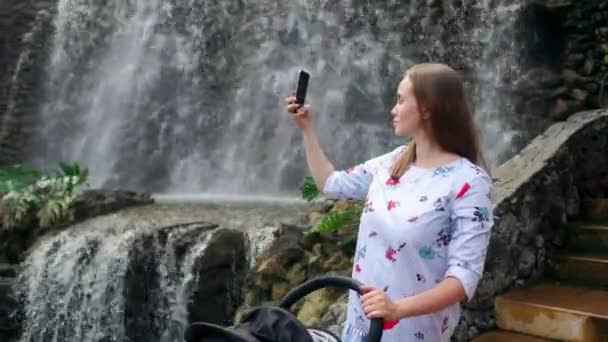 滝の近くの彼女の赤ちゃんを連れて歩いて若い母親がソーシャルネットワー キングと彼女の家族と一緒に旅行しながらオンライン投稿のスマート フォンで写真を撮る. — ストック動画