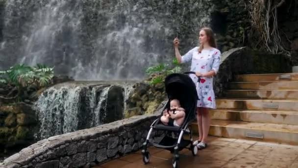 Een jonge moeder wandelen met haar baby in de buurt van de waterval neemt foto's op haar smartphone onderweg met haar familie. — Stockvideo