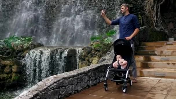 Un giovane padre che cammina con un bambino su una sedia a rotelle vicino alla cascata scatta foto sul suo smartphone per i social network e postando su Internet mentre viaggia con la sua famiglia . — Video Stock