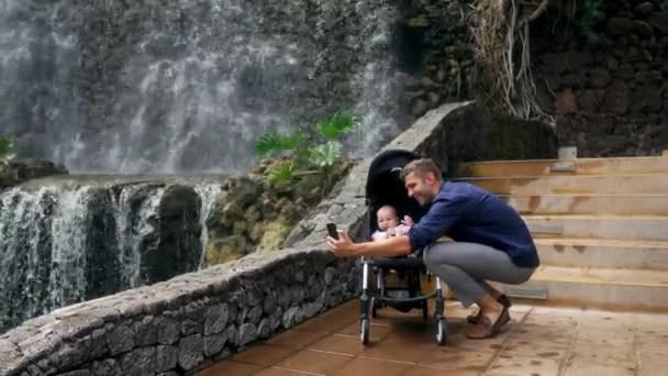 Un giovane padre che cammina con un bambino su una sedia a rotelle vicino alla cascata scatta foto sul suo smartphone per i social network e postando su Internet mentre viaggia con la sua famiglia. Il concetto di — Video Stock