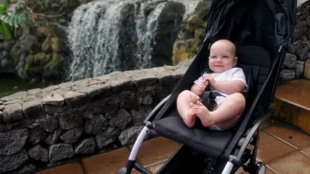 The baby sits in a wheelchair near the waterfall and laughs looking at the camera — Stock Video