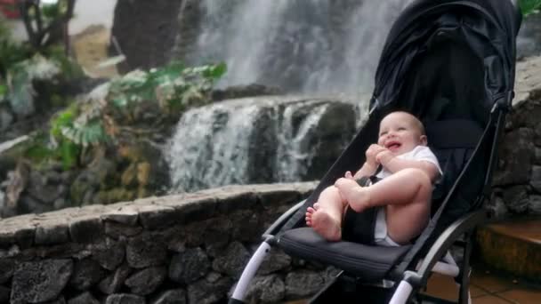 Il bambino si siede su una sedia a rotelle vicino alla cascata e ride guardando la fotocamera nella remsa viaggiare con i genitori sulle Isole Canarie . — Video Stock