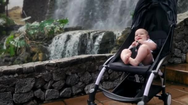 Barnet sitter i rullstol nära vattenfallet och skrattar tittar på kameran i remsa reser med föräldrar på Kanarieöarna. — Stockvideo