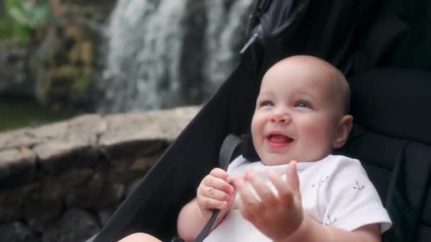 O bebê se senta em uma cadeira de rodas perto da cachoeira e ri olhando para a câmera na viagem remsa com os pais nas Ilhas Canárias . — Vídeo de Stock