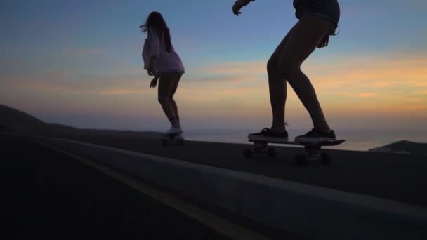 Zwei Mädchen auf Skateboards fahren bei Sonnenuntergang auf der Straße gegen Felsen und Himmel — Stockvideo