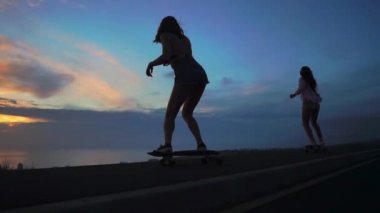Şort ve spor ayakkabısı'de iki kız kız güzel gökyüzüne yükselen güneşin karşı yamacında kaykay binmek. Ağır çekim 120 fps