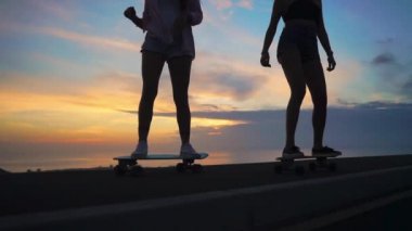 İki kız kız şort ve spor ayakkabılar bacaklar Close-up güzel gökyüzüne yükselen güneşin karşı yamacında kaykay binmek. Ağır çekim 120 fps