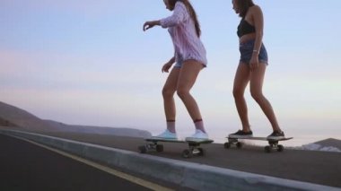 Şort ve spor ayakkabısı'de iki kız kız güzel gökyüzüne yükselen güneşin karşı yamacında kaykay binmek. Ağır çekim 120 fps