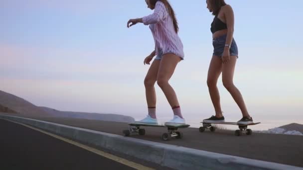 Две девушки в шортах и кроссовках катаются на скейтбордах по склону на фоне прекрасного неба восходящего солнца. Медленное движение 120 кадров в секунду — стоковое видео