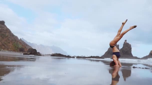 Schöne Frau am Strand des Ozeans mit schwarzem Sand führt Gymnastikübungen durch, die einen gesunden Lebensstil und Outdoor-Training praktizieren — Stockvideo