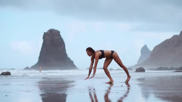 Ginasta menina na praia faz uma roda e se torna uma ponte de pose de pé em suas mãos e pés flexionando nas costas no oceano — Vídeo de Stock