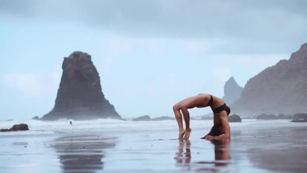 Ginasta menina na praia faz uma roda e se torna uma ponte de pose de pé em suas mãos e pés flexionando nas costas no oceano — Vídeo de Stock