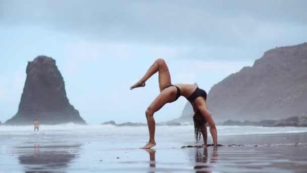 Красивая женщина на берегу океана с черным песком выполняет гимнастические упражнения, практикуя здоровый образ жизни и открытые тренировки — стоковое видео