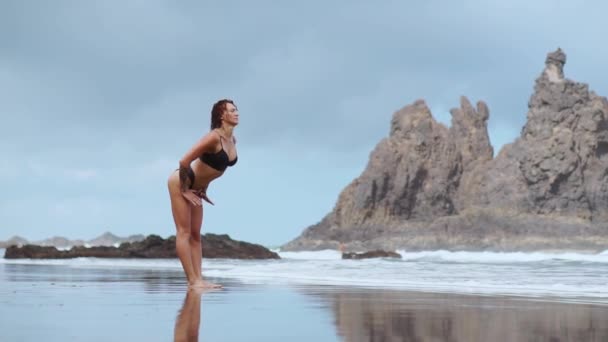 Ein junges schlankes blondes Mädchen im Badeanzug hört Musik mit großen schwarzen Kopfhörern und tanzt an einem tropischen Strand am Ufer in den aufkommenden Wellen des Ozeans — Stockvideo