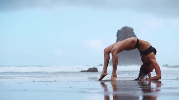 En flicka med en kort frisyr klädd i shorts och en vit tröja gör ett snöre på bakgrunden av havet och stranden. Att idrotta i naturen. Stretching, Yoga pose. — Stockvideo