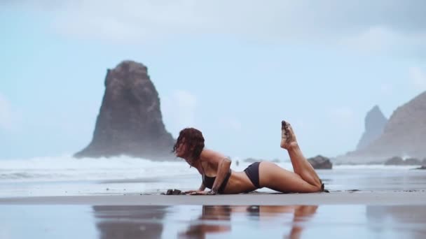 Una joven rubia esbelta en traje de baño escucha música con grandes auriculares negros y baila en una playa tropical en la orilla en las olas del océano — Vídeo de stock
