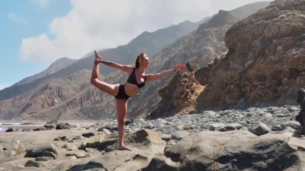 Jolie fille blonde en haut noir se détend dans la pose de yoga lotus sur tapis violet contre les vagues de l'océan qui courent sur les rochers — Video