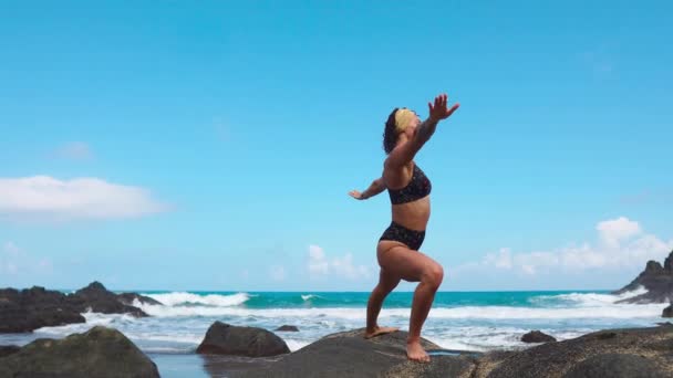 Söt blond flicka i svart topp slappnar av i yoga pose lotus på lila matta mot havsvågor som körs på stenar — Stockvideo