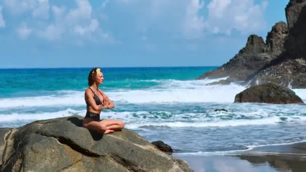 Bella ragazza bionda in top nero si rilassa in posa yoga loto sul tappetino viola contro le onde dell'oceano in esecuzione su rocce — Video Stock