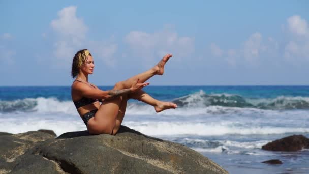 Женщина занимается фитнесом на пляже с вулканическим черным песком в купальнике, сидя на скале вокруг гор лавы. Концепция здорового образа жизни и отдыха на островах — стоковое видео