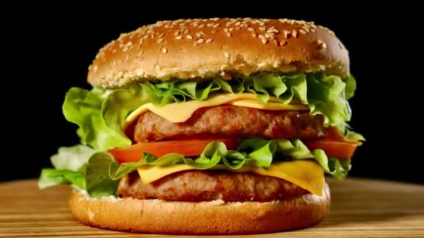 Velký Burger s hovězí kotleta, rajčaty, žampiony a okurky s rozpečeným sýrem se otočí na dřevěné desce na černém pozadí. — Stock video