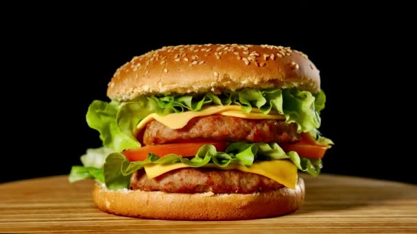 Gran hamburguesa con chuleta de res, tomates, champiñones y pepinos con queso derretido gira sobre un tablero de madera sobre fondo negro . — Vídeos de Stock