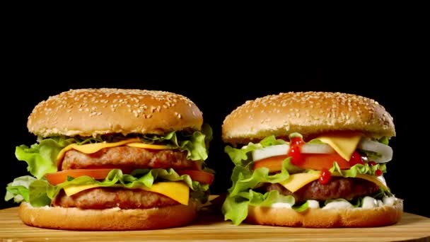 Крупный план двух аппетитных бургеров с кунжутными булочками, вращающимися на черном фоне, бесшовных петельных снимков быстрого питания  . — стоковое видео