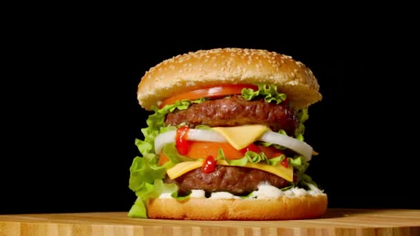 Grande hambúrguer com costeleta de carne, tomates, cogumelos e pepinos com queijo derretido gira em uma tábua de madeira no fundo preto . — Vídeo de Stock