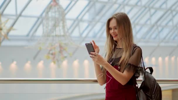 Girl with a backpack talking on video coll through headphones and a mobile phone at the airport. posing for selfie on smartphone camera, hipster girl enjoying radio broadcast making photo for picture — Stock Video