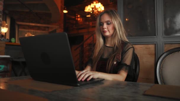 Πλαϊνή όψη. Νεαρή γυναίκα που κάθεται στο τραπέζι και σημειώσεις στο Σημειωματάριο. Στο τραπέζι είναι laptop, το smartphone και το φλιτζάνι του καφέ. Στον υπολογιστή οθόνη γραφικά και γραφήματα. Φοιτητής Μαθαίνοντας διαδικτυακά. Blogger. — Αρχείο Βίντεο