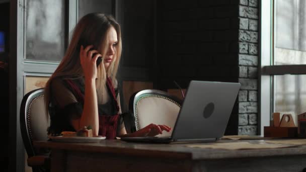 Νεαρή γυναίκα που κάθεται στο καφενείο στο ξύλινο τραπέζι, πίνοντας καφέ και χρησιμοποιώντας το smartphone. Στο τραπέζι είναι laptop. Κορίτσι περιήγηση στο internet, chatting, blogging. Θηλυκό κρατώντας τηλέφωνο και κοιτάζοντας στην οθόνη του. — Αρχείο Βίντεο