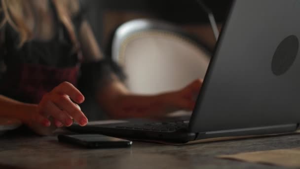 Mujer joven sentada en la cafetería en la mesa de madera, bebiendo café y usando un teléfono inteligente.En la mesa está el portátil. Chica navegando por Internet, charlando, blogueando. Mujer sosteniendo el teléfono y mirando en su pantalla . — Vídeos de Stock