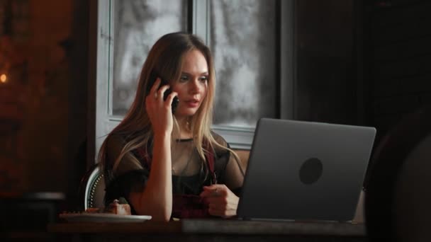 Young woman sitting in coffee shop at wooden table, drinking coffee and using smartphone.On table is laptop. Girl browsing internet, chatting, blogging. Female holding phone and looking on his screen. — Stock Video