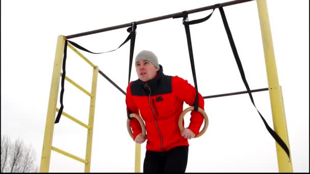 Un homme vêtu d'une veste rouge faisant de la gymnastique Stationnaire Anneaux en plein air en hiver — Video