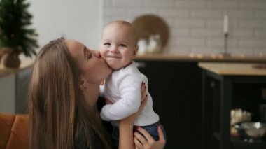 Anne Bebek ve çocuk gülümsüyor sevgili annesine bakarak sarılmak. Birlikte beyaz mutfakta Noel arifesinde çelenk ve Noel ağaçları arka plan üzerinde bekletin. Mutlu anne ve bebek