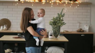 Anne Bebek ve çocuk gülümsüyor sevgili annesine bakarak sarılmak. Birlikte beyaz mutfakta Noel arifesinde çelenk ve Noel ağaçları arka plan üzerinde bekletin. Mutlu anne ve bebek