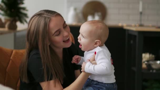 Maminka drží dítě a dětské úsměvy při pohledu na svou milovanou matku. Společně se stát v bílé kuchyni na Štědrý den na pozadí věnce a vánoční stromky. Šťastný matka a dítě — Stock video