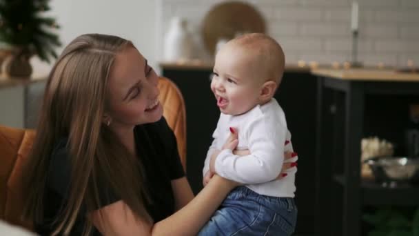 Mamãe abraça o bebê e a criança sorri olhando para sua amada mãe. Juntos ficar na cozinha branca na véspera de Natal no fundo de guirlandas e árvores de Natal. Mãe feliz e bebê — Vídeo de Stock