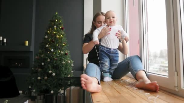 Νεαρή μητέρα κάθεται με το γιο της στο περβάζι του παραθύρου διακοσμημένα με Χριστουγεννιάτικο στεφάνι και κοιτάζοντας έξω από. Είναι μιλάμε και χαμογελαστός. Ευτυχισμένη οικογένεια στο σπίτι — Αρχείο Βίντεο