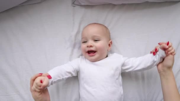 Un niño en una camiseta blanca acostado en una cama blanca mirando a la cámara y riendo en cámara lenta, vista superior — Vídeos de Stock