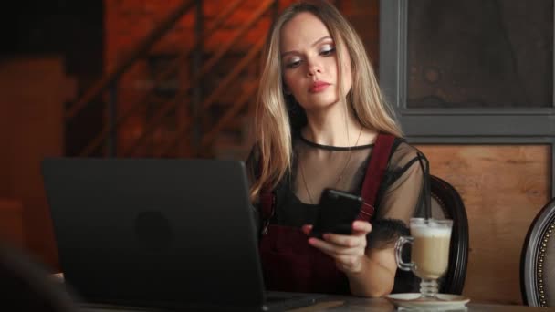 Молодая женщина сидит в кафе за деревянным столом, пьет кофе и пользуется смартфоном. На столе ноутбук. Девушки просматривают интернет, общаются, ведут блог. Женщина держит телефон и смотрит на его экран . — стоковое видео