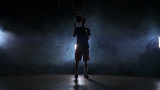 Un homme sous forme sportive tourne un ballon de basket sur son doigt en effectuant un tour en regardant la caméra sur le terrain de basket-ball autour de la fumée au ralenti . — Video