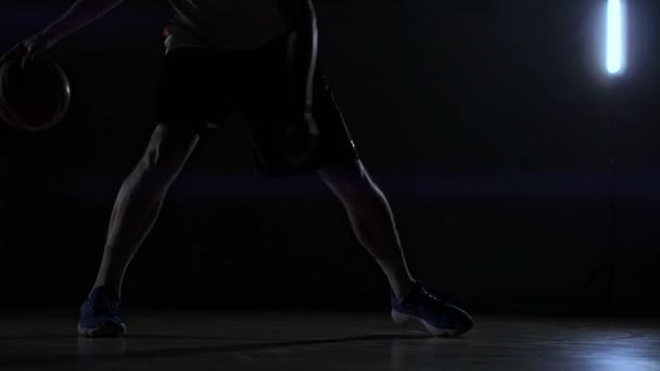 Чоловік з баскетболом на темному баскетбольному майданчику на тлі баскетбольного кільця в диму показує навички дриблінгу, освітлені трьома ліхтарями під час підсвічування — стокове відео