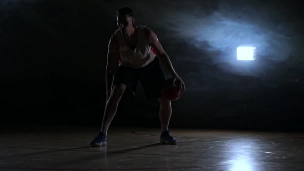 Один молодий дорослий чоловік, баскетболіст смикає м'яч, темний в приміщенні баскетбольний майданчик повільний рух — стокове відео