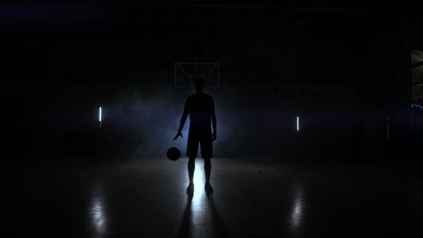 Баскетболист отбивает мяч в свете светильников, сияющих сзади, стоит в Думе и в замедленной съемке стучит мячом по полу в баскетбольном зале. — стоковое видео
