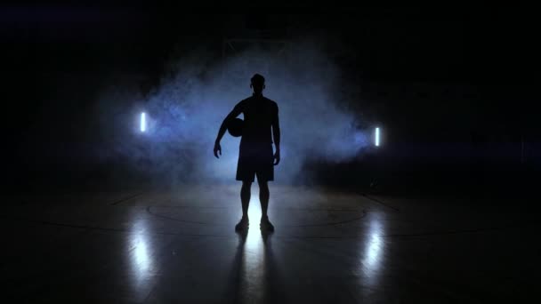Basketball joueur frappe la balle à la lumière des lampes qui brille derrière se tient dans la Douma et frappe la balle sur le sol dans la salle de basket au ralenti — Video