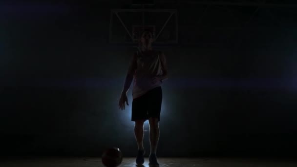 Koszykarz stoi na plac zabaw ciemny i posiada piłkę w ręce i wygląda w aparat w ciemności z podświetleniem w zwolnionym tempie i okolice: dym — Wideo stockowe