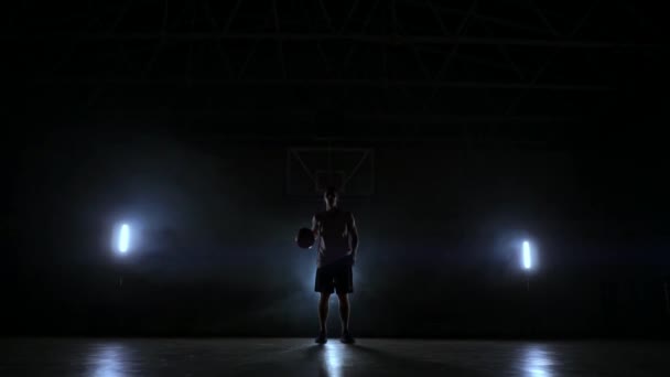 Basketspelare står på en dark Playground och håller bollen i händerna och ser in i kameran i mörkret med en bakgrundsbelyst i slow motion och runt rök — Stockvideo