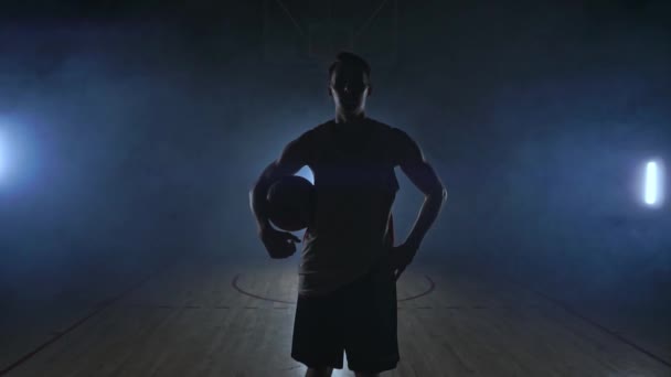 O jogador de basquete vai para a câmera e bate a bola no chão, em seguida, pára e segura a bola olhando para a câmera — Vídeo de Stock
